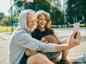 Älterer Mann sitzt mit Kind auf dem Basketballfeld und beide lächeln in die Handykamera