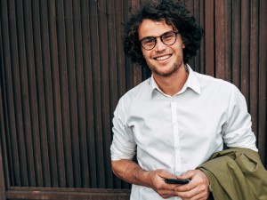 Junger Mann mit Brille hält sein Handy in der Hand und lächelt in die Kamera