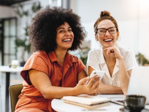 Zwei Frauen sitzen zusammen an einem Tisch und lachen in die Kamera