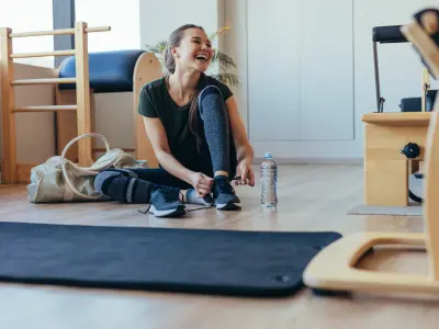 Eine junge Frau sitzt lächelnd in einem Fitnessraum und bindet sich die Schuhe zu.