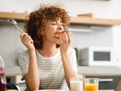Eine junge Frau sitzt am Küchentisch und beißt in ein Brot mit Marmelade.