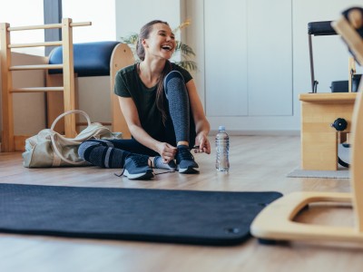 Eine junge Frau sitzt lächelnd in einem Fitnessraum und bindet sich die Schuhe zu.