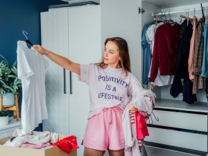 Ausmisten: Junge Frau steht vor Kleiderschrank und schaut Klamotten an