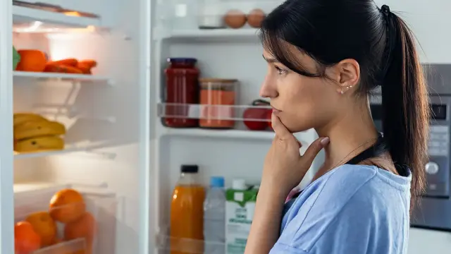 Junge Frau steht vor offenem Kühlschrank und überlegt.