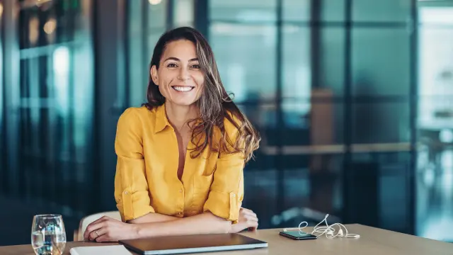 Frau sitzt mit Laptop am Tisch und lächelt in die Kamera