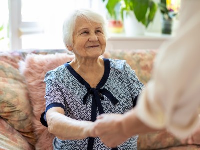 Eine ältere Frau sitzt auf einem Sofa. Vor ihr steht eine Person und hält ihre Hände fest.