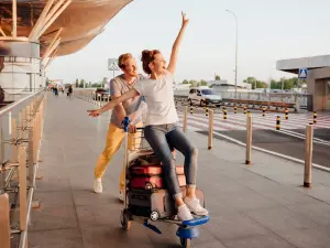 Junger Mann schiebt eine junge, gut gelaunte Frau auf einem Kofferwagen vor einem Flughafengebäude