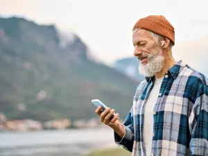 Älterer Mann mit Kopfhörern und Handy an einem Bergsee